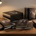 Plusieurs éditions de romans graphiques de fantasy sombre sont disposées sur une table en bois, avec des formats reliés, brochés et grand format éclairés par une lampe de bureau.
